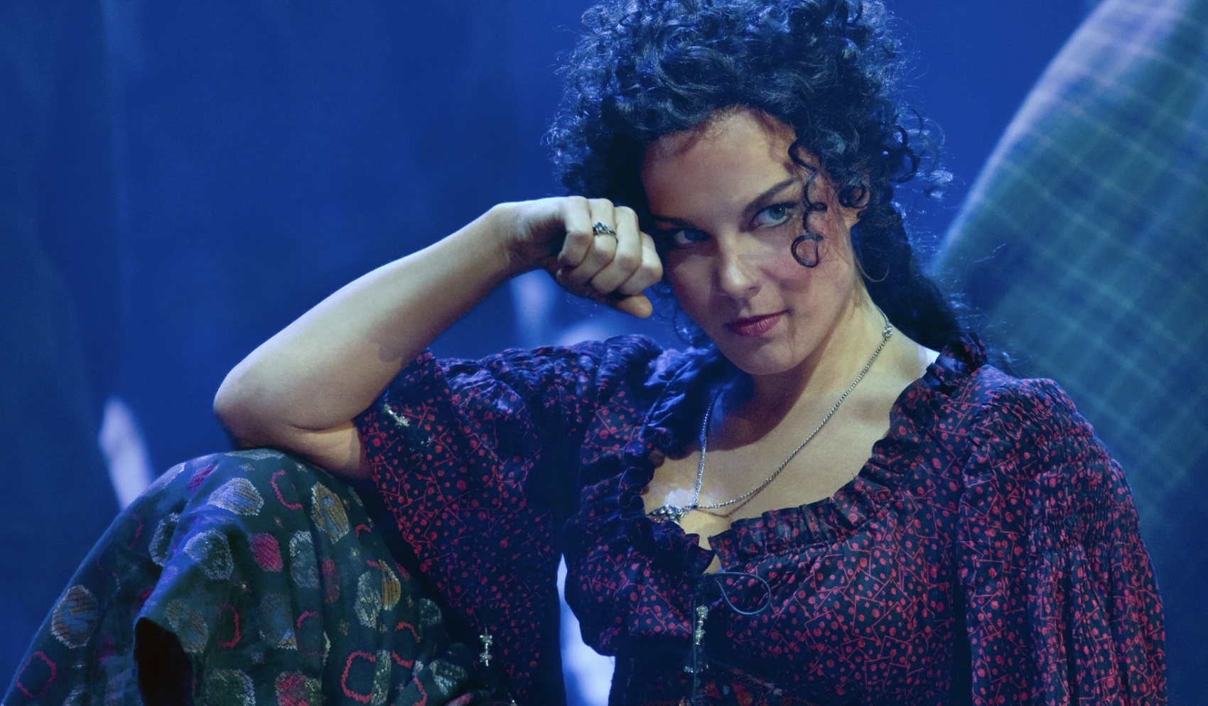 Actress in stage performance of Carmen sits with her elbow on a bent knee and her head resting on her fist